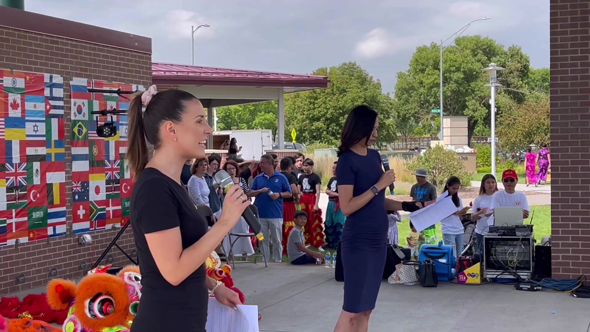 Two event hosts speaking outdoors with community members and international flags behind them